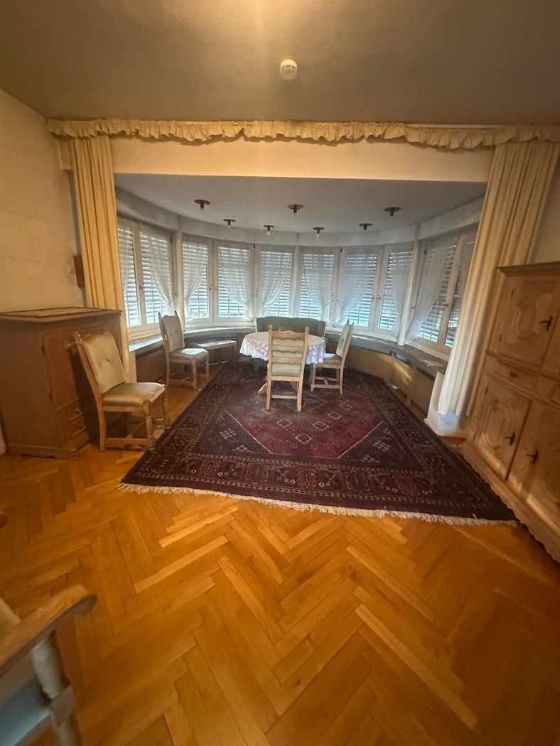 Wohnzimmer Freckenhorst vor der Entrümpelung – vollständig eingerichteter Raum mit Holzschränken, Stühlen, großem Teppich und Vorhängen im Rundbogen-Bereich